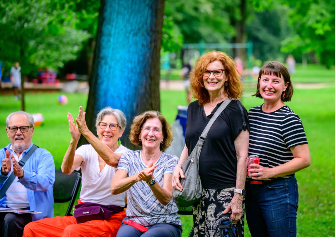 Photo of a group of people, some seated and some standing, in a park. Those seated are clapping.
