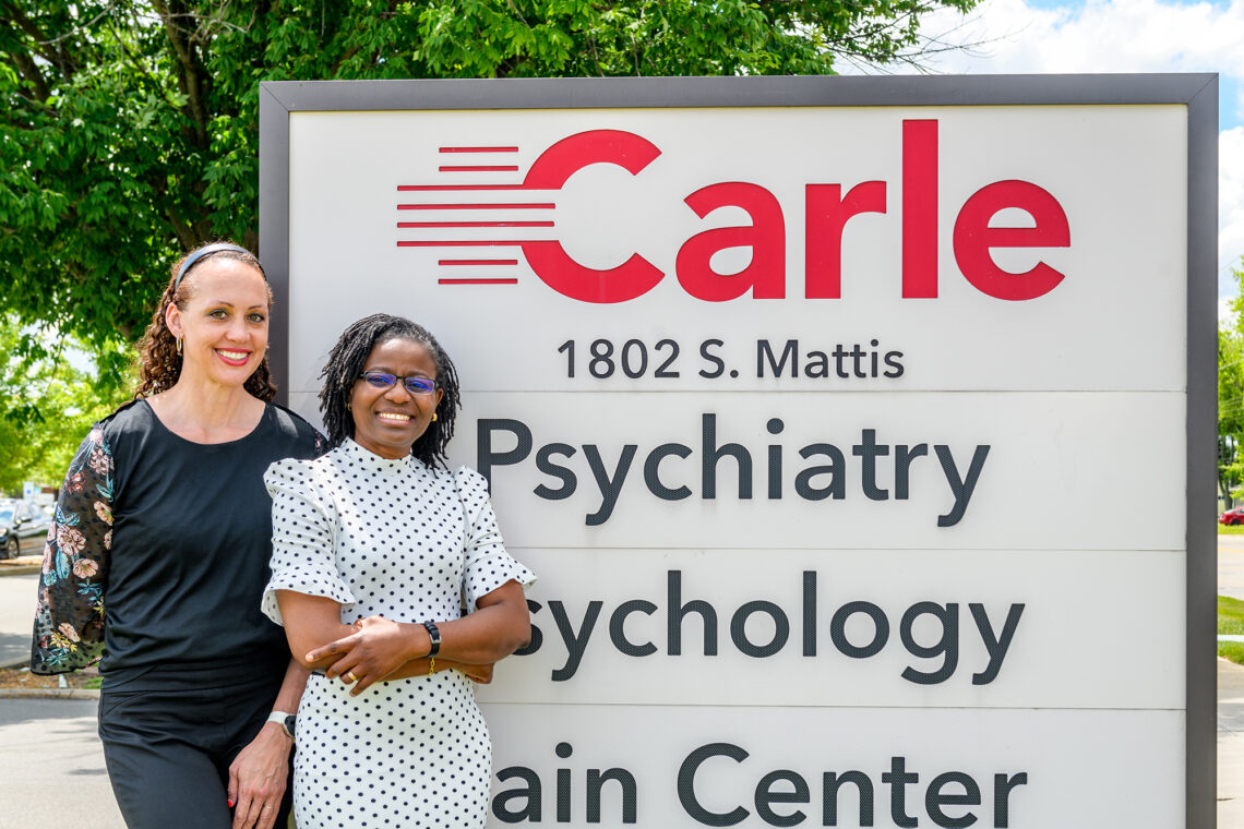 Social work professor Karen Tabb and psychiatrist Dr. Mercy Eigbike standing outdoors in front of a Carle sign.