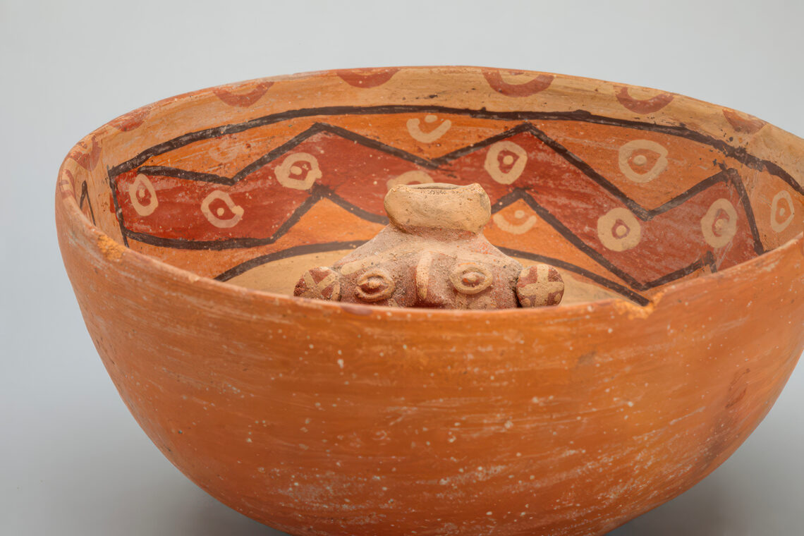 Photo of an orange-colored ceramic ceremonial bowl with a ceramic figure inside seen peeking over the rim of the bowl, from Peru.