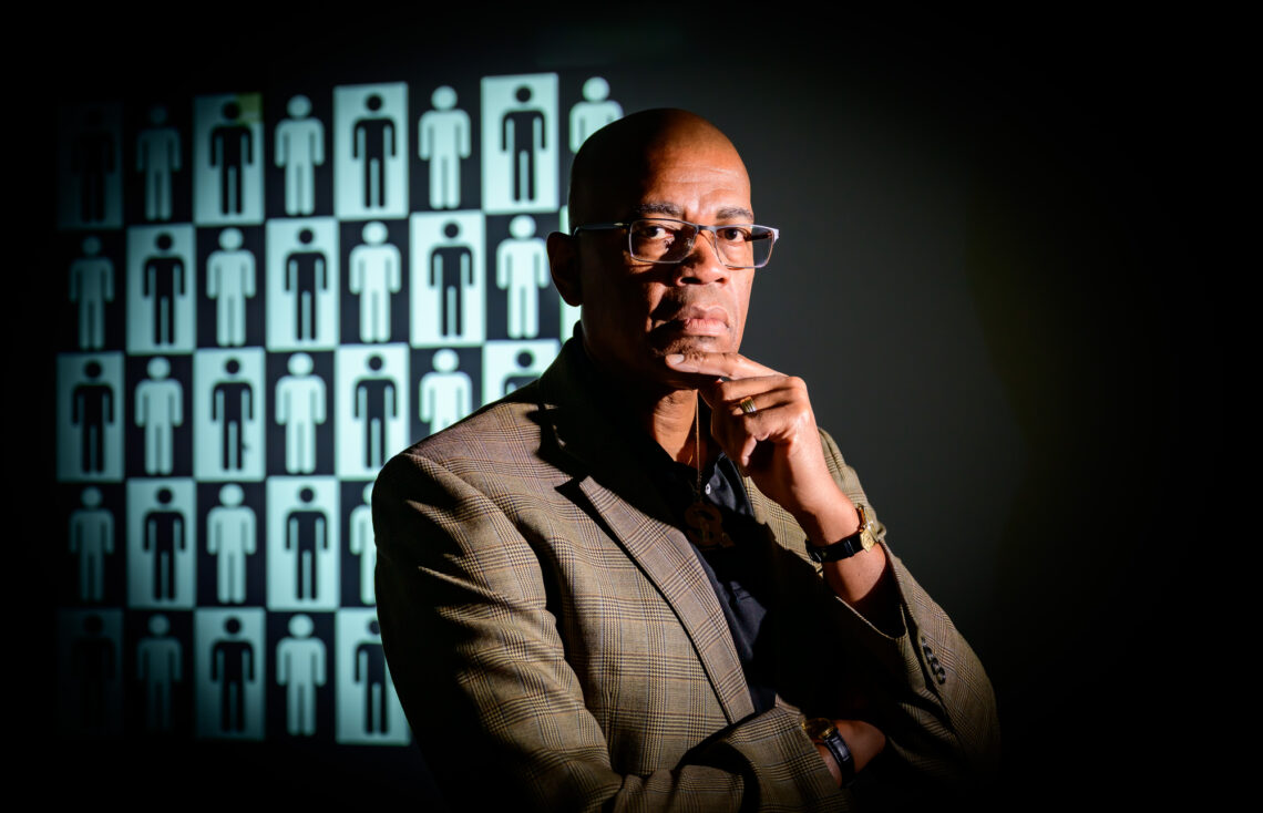 Reuben May wearing a gray suit jacket and black shirt in a thoughtful pose with one finger across his chin