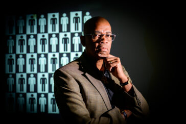 Reuben May wearing a gray suit jacket and black shirt in a thoughtful pose with one finger across his chin