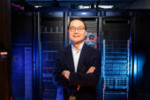 A man wearing a dark suit stands with arms crossed in front of a server bank