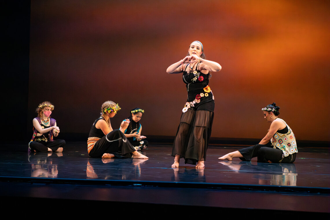 Photo of several dancers onstage, most of them sitting on the floor and one standing. They have flower crowns on their heads and flowers on their costumes.