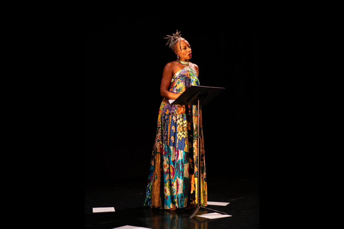 Photo of Cynthia Oliver wearing a long, colorful dress, standing in front of a music stand with papers on the floor at her feet.