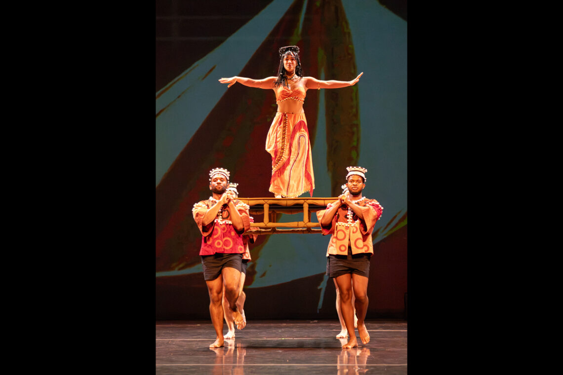 Dancers onstage, with men carrying a platform where a woman stands with outstretched arms.