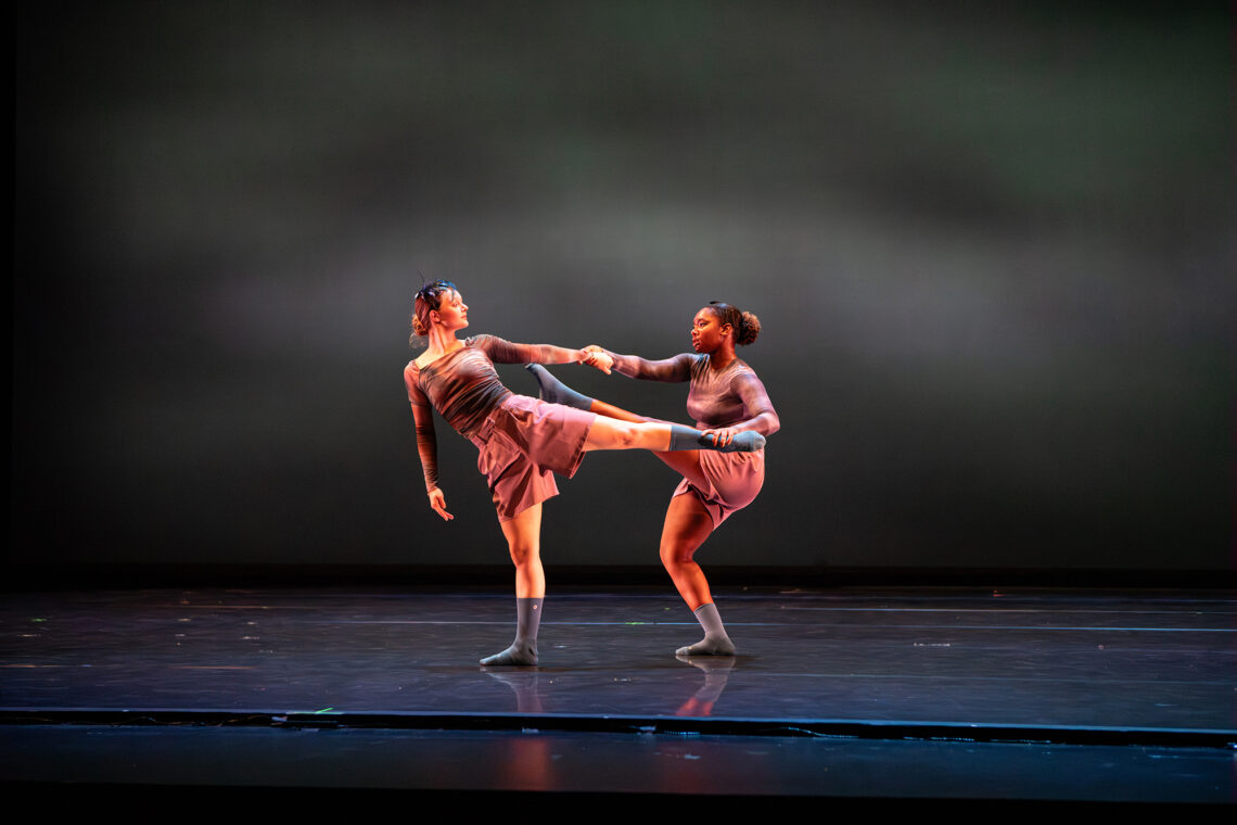 Photo of two dancers onstage, each with one leg and arm extended toward the other and holding hands with their extended arms.