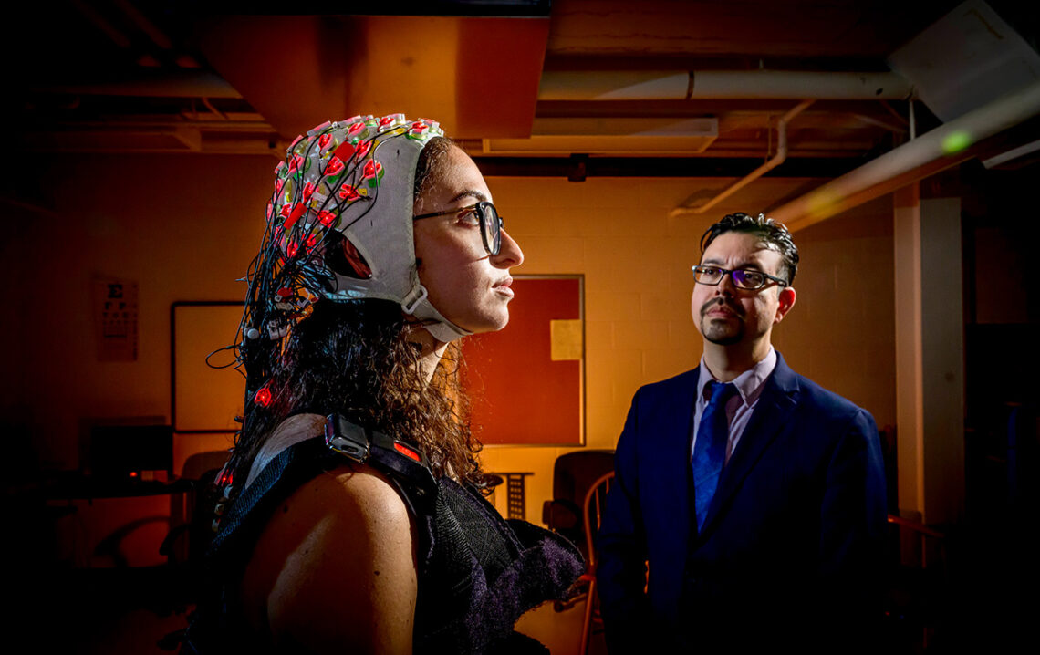 Participant stands looking up and to the right, her face illuminated. She is wearing a smart shirt, which looks like a thick vest, and an electrode-studded cap. The individual electrodes are glowing bright red. Hernandez stands to the right, observing the setup.