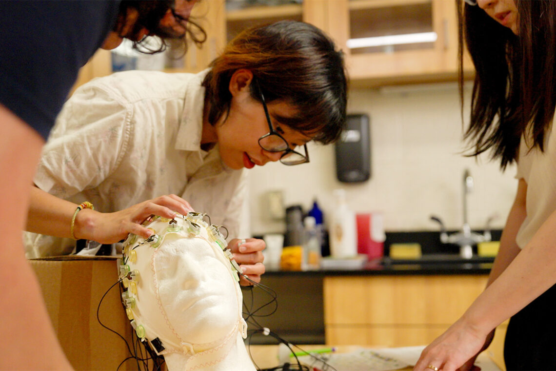 Student looks closely at the electrode-studded cap, which is on a styrofoam wig-head.