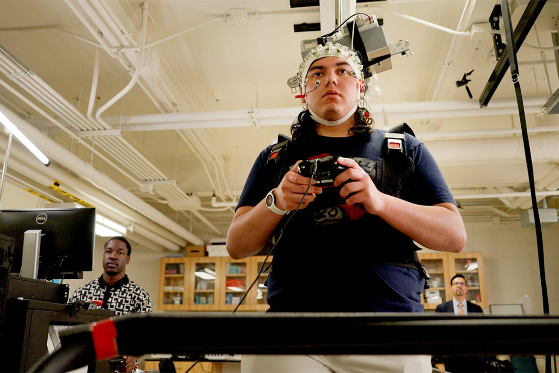 The participant stands on a treadmill facing the camera and holding a controller that allows him to quickly respond to a stimulus on a computer monitor.