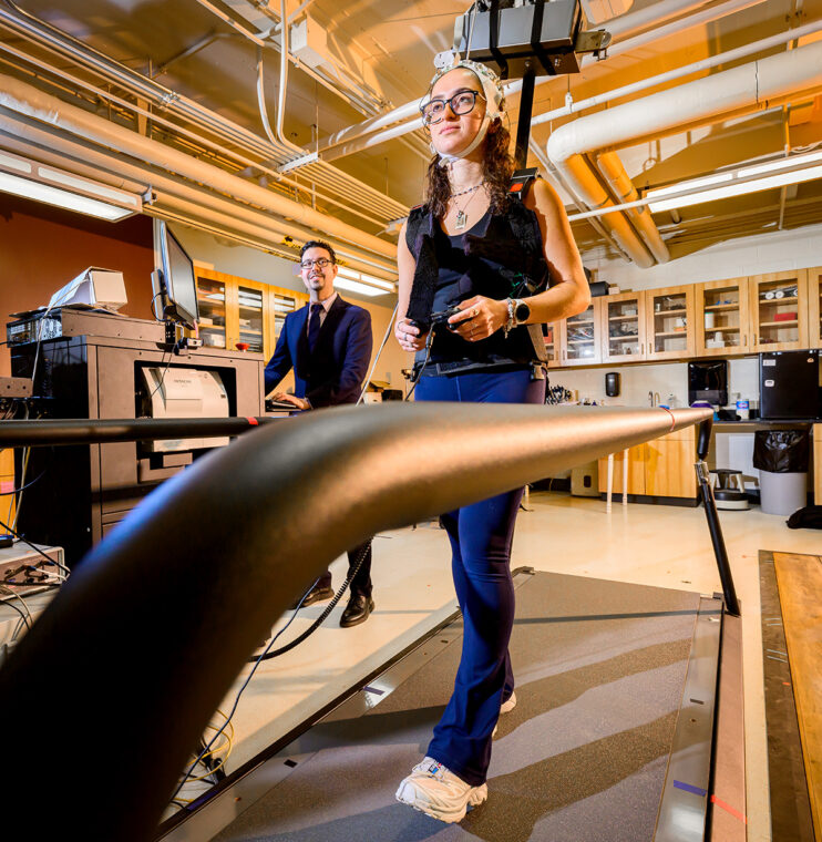 Hernandez stands behind a participant who is geared up with wearable technology and walking on a treadmill with a controller in her hands.