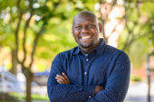 Moses Okumu standing outside wearing a blue shirt with green trees in the background.