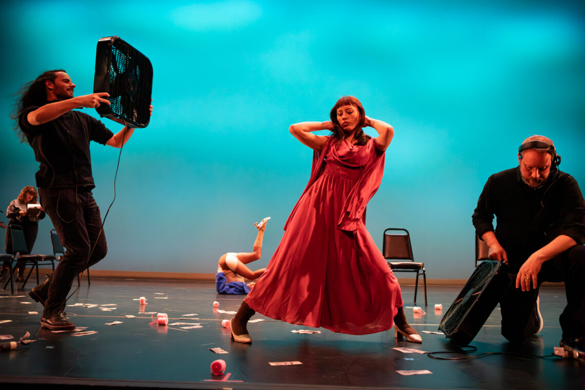 Photo of a woman in a long, pink-colored dress posing onstage with a man aiming a box fan at her.