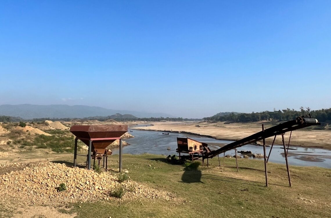 A photograph of an abandoned gravel machine sits in disuse on the riverbank, with a pile of leftover stones, as the river in the background slowly returns to its natural form.