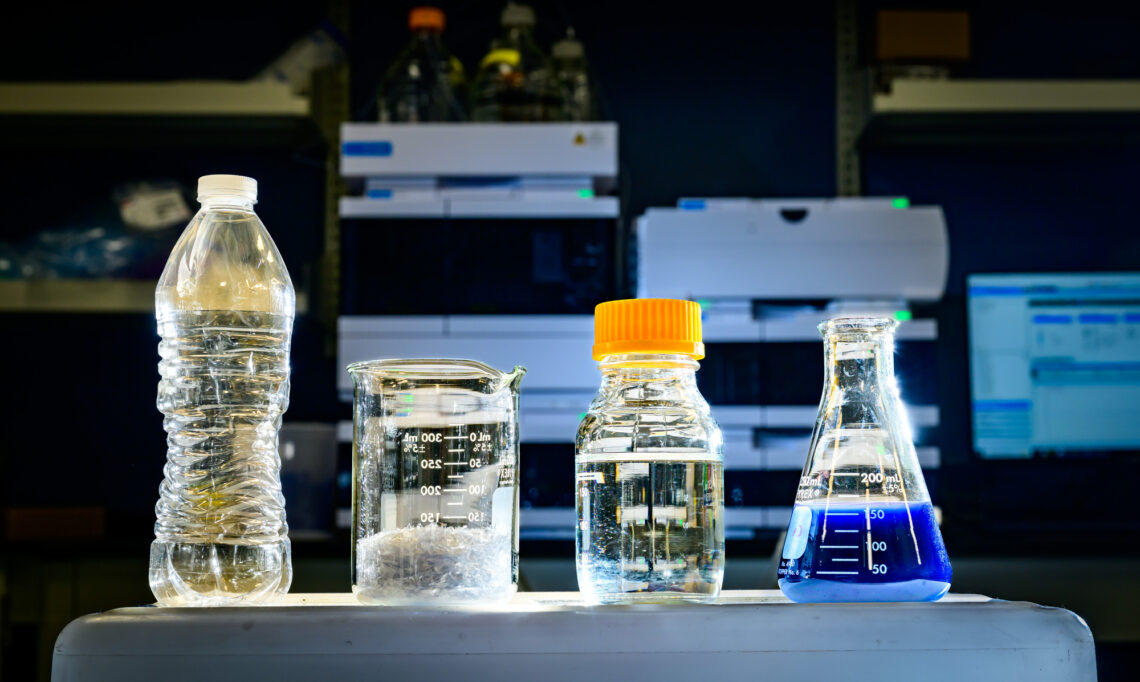 Photo from the lab of four containers. 1) a plastic bottle, 2) a beaker with shredded plastic, 3) a capped bottle with pyruvate and 4) a flask with blue dye.