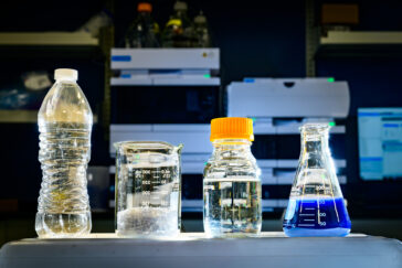 Photo from the lab of four containers. 1) a plastic bottle, 2) a beaker with shredded plastic, 3) a capped bottle with pyruvate and 4) a flask with blue dye.