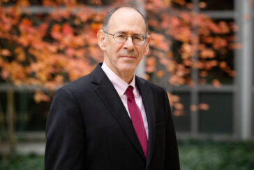 A man in a dark suit and glasses, with orange foliage in the background