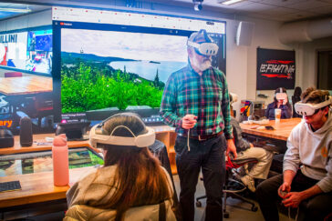 Photo of Robert Pahre wearing virtual reality goggles in front of a screen with a wilderness photo and surrounded by seated students also wearing VR goggles.