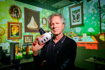 Portrait of John Toenjes holding virtual reality goggles, with an image from his VR game "Master Dancer" in the background.