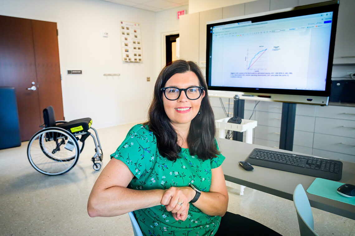 Health and kinesiology professor Laura Rice seated before a large monitor and keyboard with a wheelchair in the background.