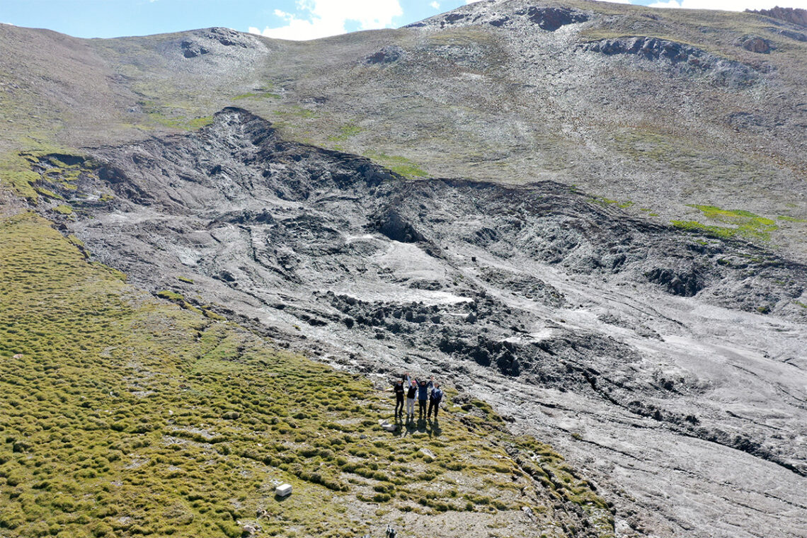 Photo of a large slump on a hillside or mountainside. Four people stand on one edge of the slump, giving a sense of perspective. The slump appears to be 2-4 times deeper than their heights. 