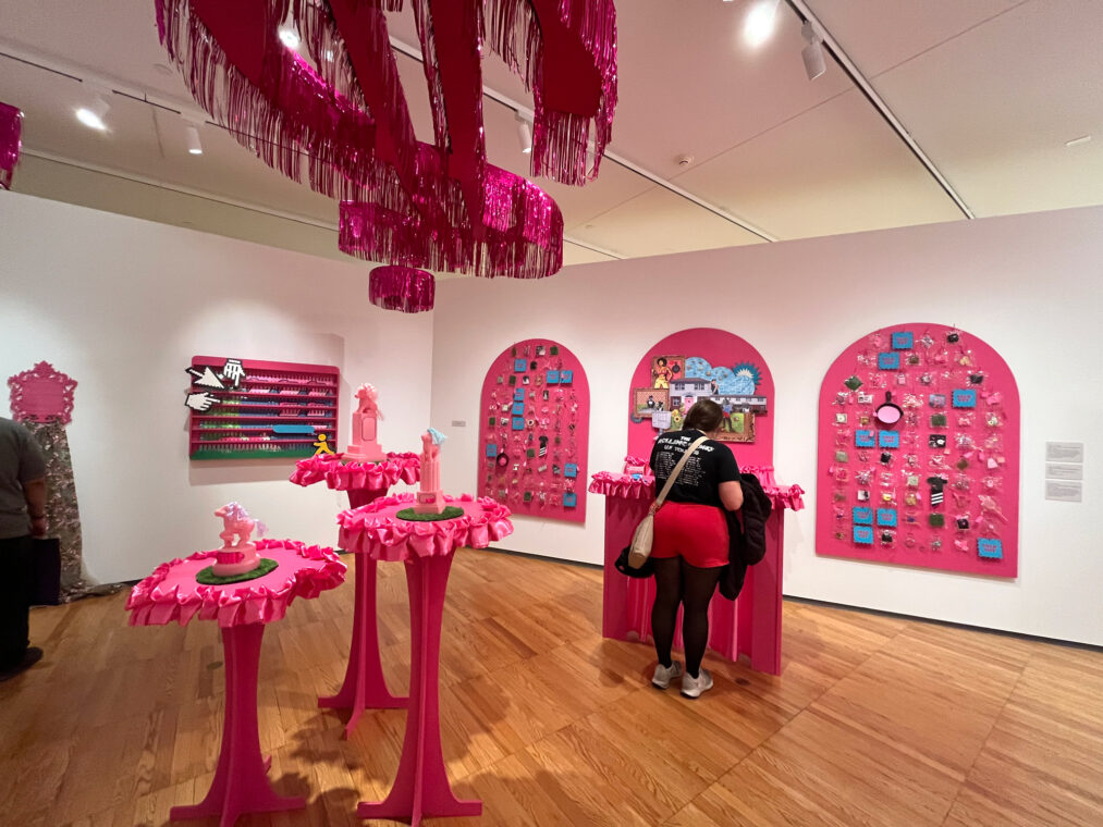 Photo of a pink installation with pink tables topped with plastic toys, pink panels on the walls with plastic bags filled with items and pink streamers hanging from the ceiling.