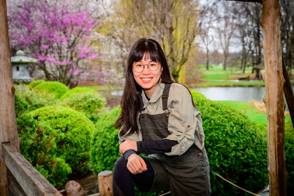 Yue (Darcy) Lu standing with one elbow on her knee with flowering spring trees in the background.