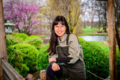 Yue (Darcy) Lu standing with one elbow on her knee with flowering spring trees in the background.