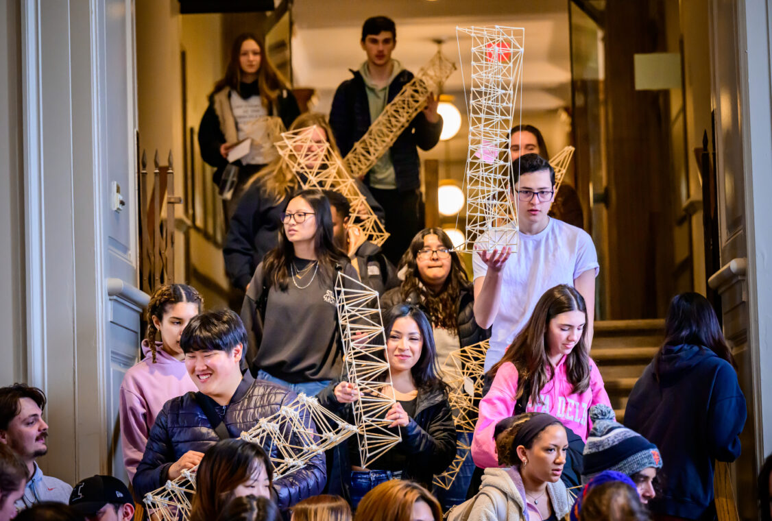 Photo of students carrying trusses made from bamboo sticks walk down a stairway into a room.