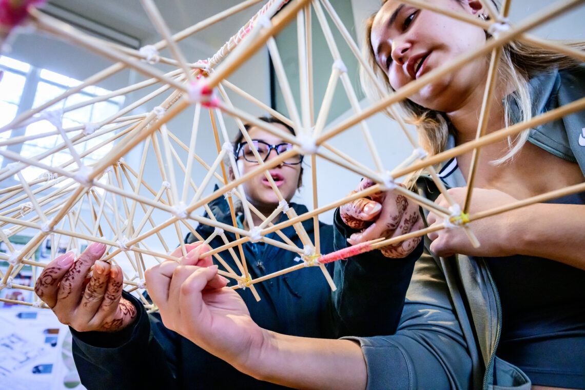 Photo of two students, shot from underneath looking up through their truss, examining the truss that they built.