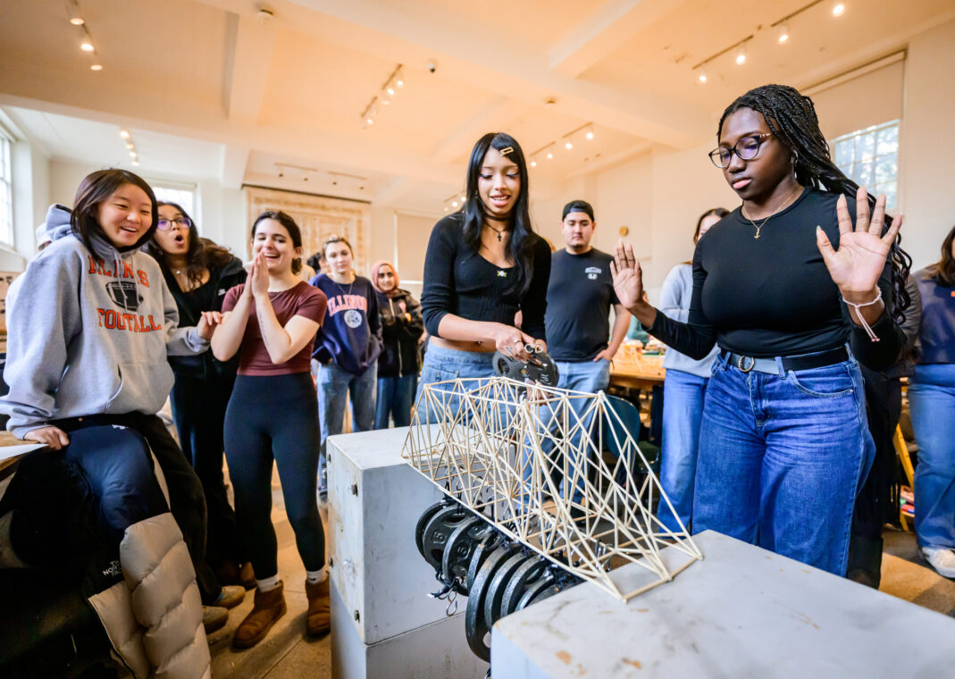Photo of students surrounding a truss made from bamboo sticks that is holding weights suspended from its bottom supports.