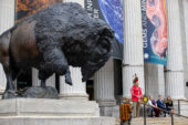 Photo of an immense bronze bison statue looming over a Native American speaker and others seated beside him outside the National Museum of Natural History.