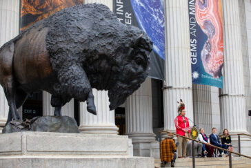 Photo of an immense bronze bison statue looming over a Native American speaker and others seated beside him outside the National Museum of Natural History.