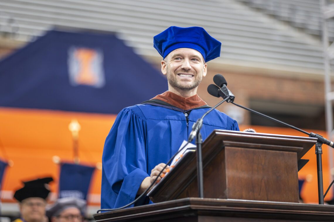 A man, Sean Evans, in graduation regalia delivering a Commencement speech