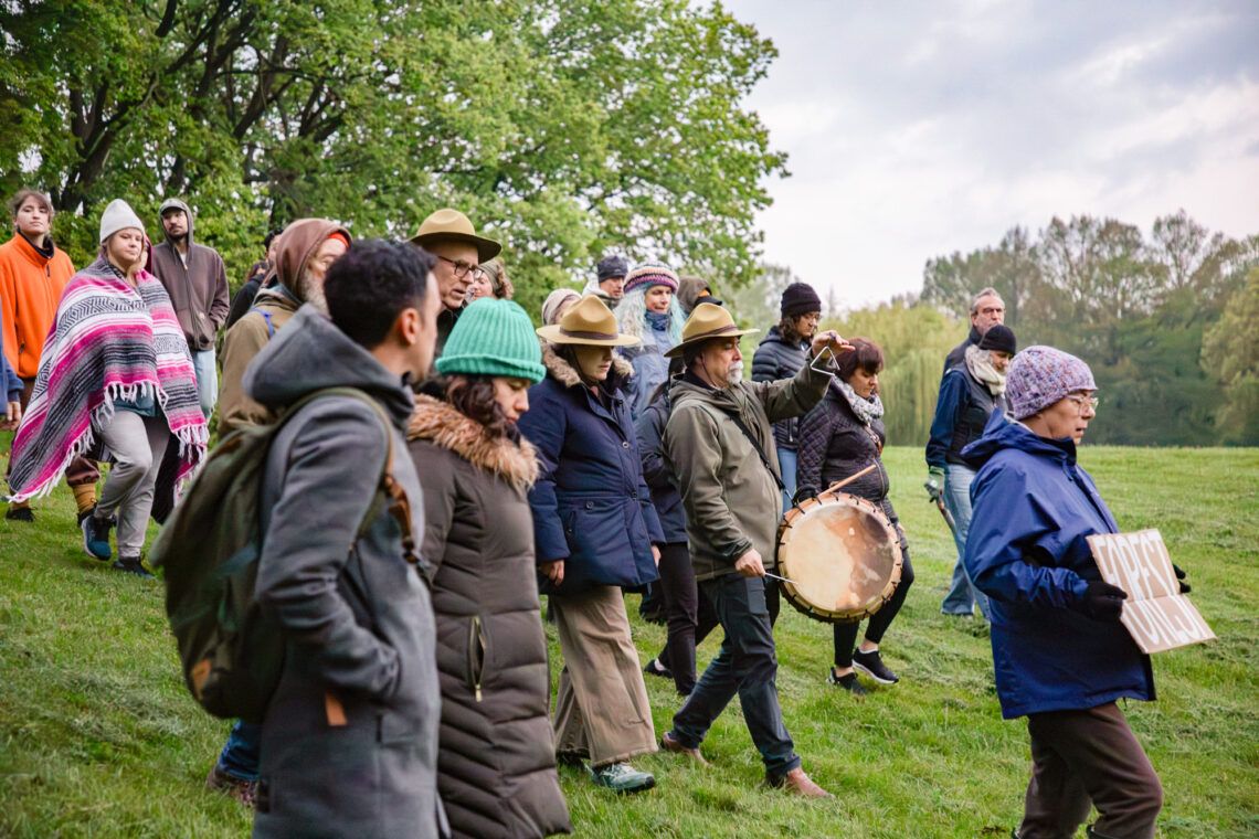 Photo of a group of people walking down a grassy hill, with one woman holding a sign that says "Forest" and a man holding a drum.
