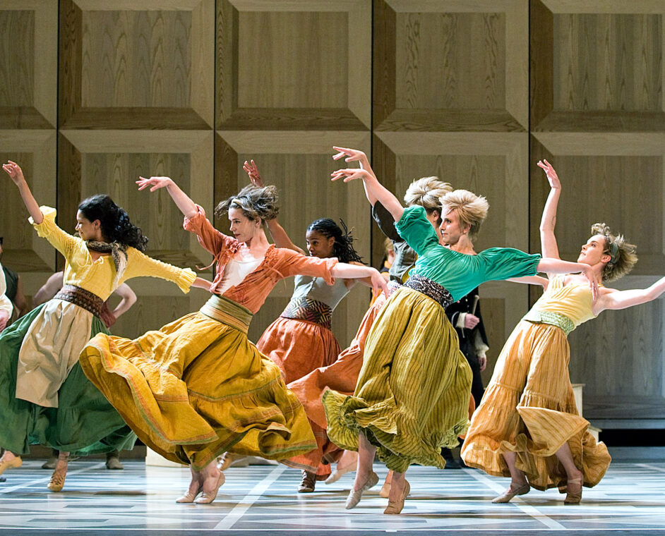 Photo of dancers from the Mark Morris Dance Group dancing onstage with their bodies angled back and their arms in the air, wearing long flowing skirts.