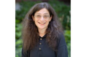 Portrait photo of May Berenbaum. She is standing outside, smiling, wearing glasses and a black top.