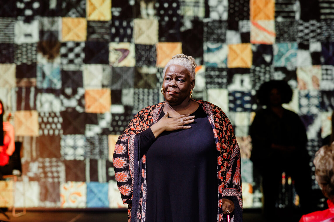 Photo of Lisa Gaye Dixon standing with her hand on her heart in front of a quilt backdrop.