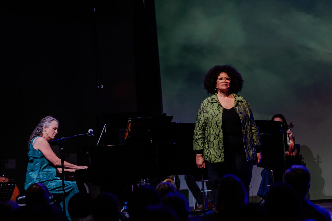 Photo of Julie Gunn seated at a piano and Adrienne Danrich standing to perform.