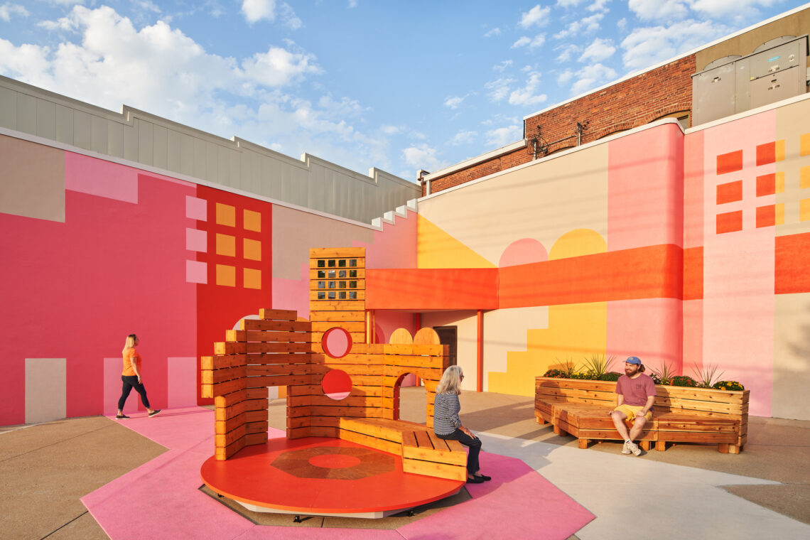 Image of an area painted in pinks, oranges and yellows with a circular wooden sitting area and another wooden bench.