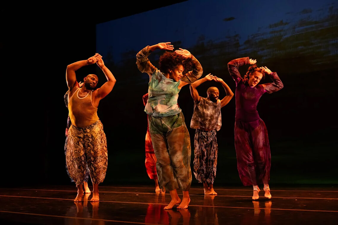 Photo of several dancers on stage with their arms above their heads, all looking down and to their left.