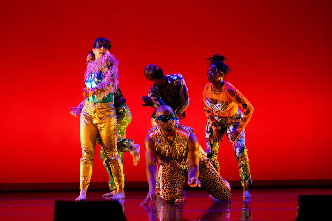 Photo of several dancers in colorful costumes in front of a red background, including a man in his knees, wearing leopard-print pants and a mask in the foreground.
