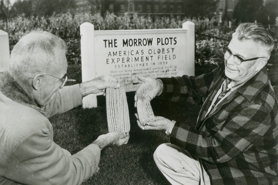 Black-and-white photo of two men each holding an ear of corn in front of a sign for the Morrow Plots.