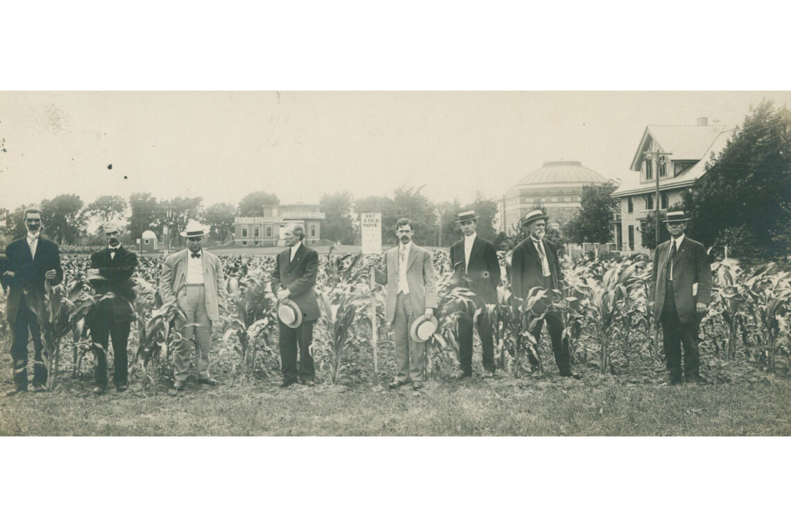 Old photo of a group of men wearing dress clothes and hats and standing in front of corn in the Morrow Plots.