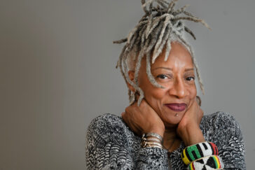Photo of Cynthia Oliver wearing a black and white sweater and beaded bracelets in African colors, with gray dreadlocks piled up on her head, and her chin in her hands.